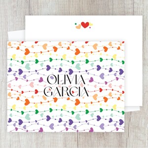 Peut inclure: Ensemble de papeterie avec une carte et une enveloppe blanches, orné d'un motif de cœurs colorés. La carte présente un motif répété de cœurs aux couleurs de l'arc-en-ciel et le nom "OLIVIA GARCIA" en noir. L'enveloppe a un petit motif de cœur.