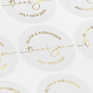 May include: White round stickers with gold lettering. The stickers say "Olivia & Alexander thank you July 10th 2024".