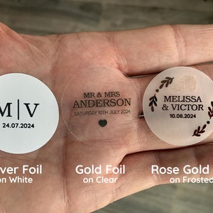 May include: Three round wedding favor stickers with different foil finishes. The first sticker is silver foil on white with the initials "M|V" and the date "24.07.2024". The second sticker is gold foil on clear with the text "MR & MRS ANDERSON" and the date "SATURDAY 10TH JULY 2024". The third sticker is rose gold foil on frosted with the text "MELISSA & VICTOR" and the date "10.08.2024".