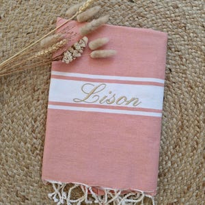 Peut inclure: Un textile rose poudré avec une bande horizontale blanche portant le nom Lison brodé en écriture cursive. Le tissu rectangulaire présente des franges blanches nouées sur le bord inférieur et est décoré d'un petit bouquet de tiges de blé séchées.