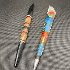 May include: Two decorative pens with colourful designs. One pen has a black base with a floral pattern and the other has a clear base with a multi-coloured pattern and a red flower at the top.