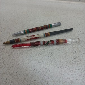 Puede incluir: Tres bolígrafos con diseños diferentes. El bolígrafo superior es plateado con rayas rojas, verdes y doradas. El bolígrafo del medio es negro con rayas blancas y doradas. El bolígrafo inferior es rojo con rayas blancas y verdes y un tubo de vidrio transparente.