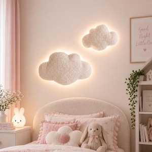 May include: Two off-white, textured, cloud-shaped wall lamps with integrated backlighting mounted on a wall above a bed. The lamps feature a soft, fuzzy surface texture and emit a warm, diffused glow around their edges, creating a halo effect.