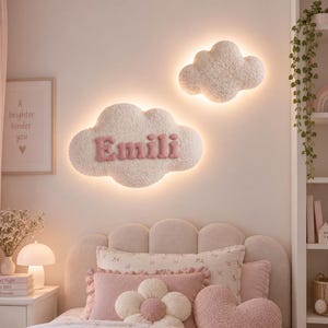 May include: Two cloud-shaped wall lights with a textured, off-white finish. The larger cloud features the name Emili in raised, pink, fabric-covered letters. Both lights emit a soft, warm glow from behind their edges, mounted on a wall.