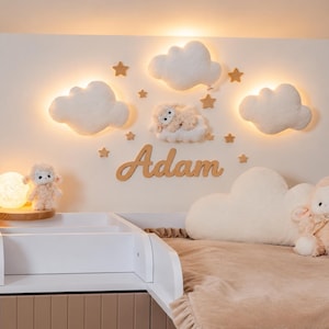 May include: A nursery wall decor set featuring three illuminated white cloud-shaped wall lights, several small wooden star cutouts, and a wooden script name sign reading Adam. Two plush sheep toys and a large cloud pillow are also included.