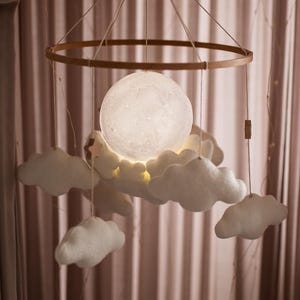 May include: A baby mobile featuring a glowing moon and fluffy white clouds. The moon is the central focus, emitting a soft light, surrounded by felt clouds. The mobile hangs from a wooden ring and is suspended by strings.