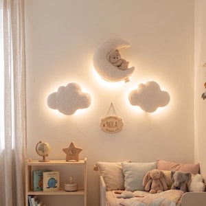 Può includere: Una cameretta con luci a parete a forma di luna e nuvole, una mensola in legno con libri e decorazioni e una culla con biancheria da letto. Un cartello di legno recita "Mila". La stanza ha una palette di colori tenui e neutri.