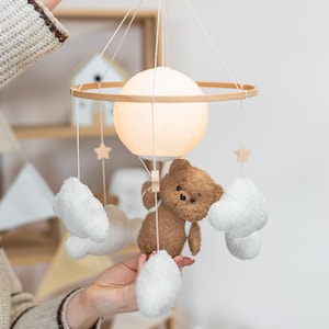 Könnte beinhalten: Baby-Mobile mit einem braunen Teddybären, weißen Wolken und Sternen. Das Mobile hat einen Holzring und ein leuchtendes kugelförmiges Licht. Eine bezaubernde Dekoration für das Kinderzimmer.