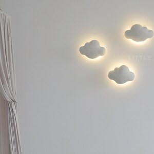 May include: Three cloud-shaped wall lights with a soft, glowing effect. The lights are a light gray color and are mounted on a white wall. The lights are arranged in a cluster, creating a whimsical and inviting atmosphere.