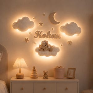 May include: A wall-mounted nursery decor set featuring two illuminated cloud-shaped lights, a crescent moon, several small stars, and a wooden cutout name sign reading Kohan. A plush bear wearing a nightcap rests on a third illuminated cloud light.