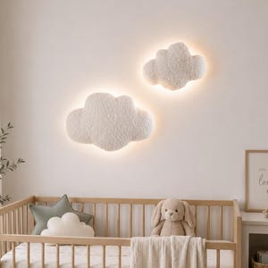 Könnte beinhalten: Zwei cremefarbene, texturierte Wandleuchten in Wolkenform mit integrierter Hintergrundbeleuchtung über einem hellen Holzbettchen. Das Bettchen enthält ein salbeigrünes Sternkissen, ein kleines Wolkenkissen und ein Plüschhasen-Spielzeug.