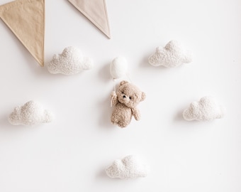 May include: A plush teddy bear holding a white balloon, surrounded by fluffy white cloud decorations on a white wall. Beige fabric pennants hang from the top. A whimsical nursery or children's room theme.