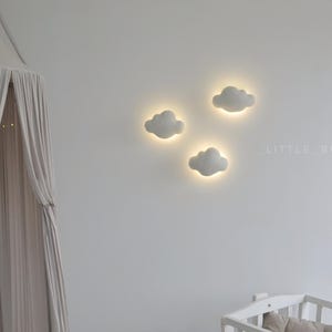 May include: Three white cloud-shaped wall lights with a soft glow, mounted on a white wall. A canopy with draped fabric hangs on the left. A crib is visible in the lower right corner. The text "LITTLE_BUNNY.CO" is on the wall.