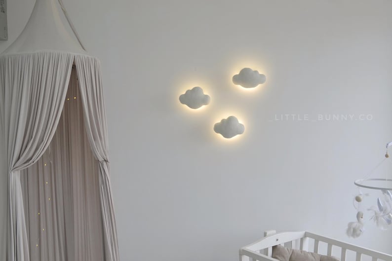 May include: Three white cloud-shaped wall lights with a soft glow, mounted on a white wall. A canopy with draped fabric hangs on the left. A crib is visible in the lower right corner. The text "LITTLE_BUNNY.CO" is on the wall.