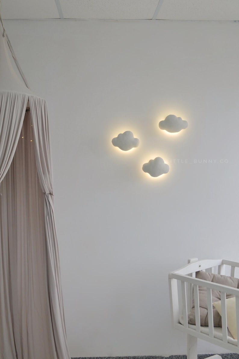 May include: Three cloud-shaped wall lights with a warm glow are mounted on a white wall. A canopy with a draped fabric hangs on the left, and a white crib with pillows is in the lower right. The website LITTLE_BUNNY.CO is visible.
