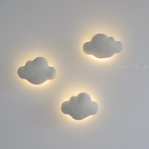 May include: Three cloud-shaped wall lights with a soft, glowing effect. The lights are white and have a fluffy texture, with a warm, yellow glow emanating from behind each cloud. The lights are mounted on a white wall.