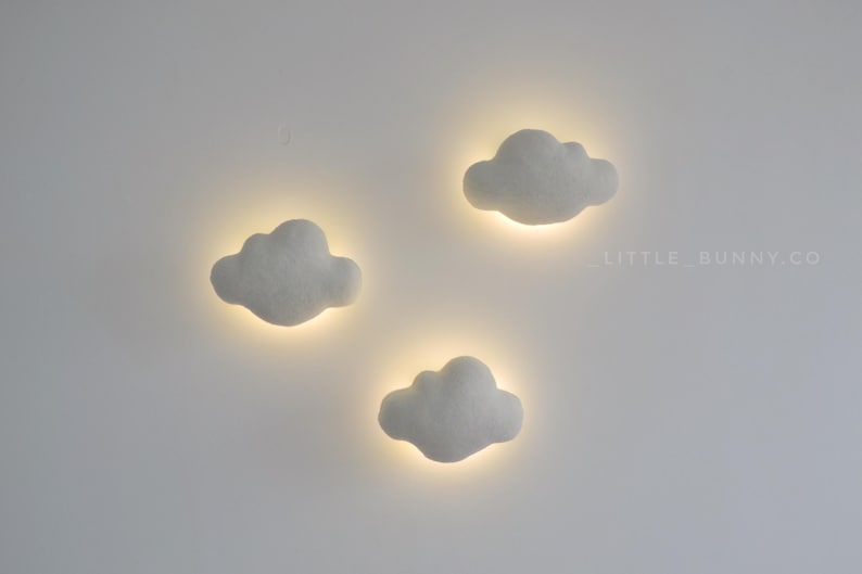 May include: Three cloud-shaped wall lights with a soft, glowing effect. The lights are white and have a fluffy texture, with a warm, yellow glow emanating from behind each cloud. The lights are mounted on a white wall.