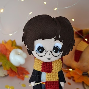 Puede incluir: Un muñeco de Harry Potter de fieltro con una bufanda roja y amarilla y sosteniendo una varita mágica. El muñeco tiene el pelo castaño, los ojos negros y lleva una túnica negra.