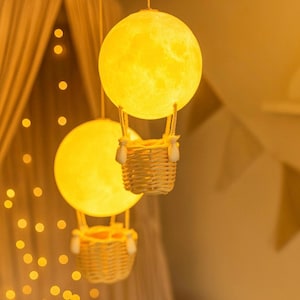 Puede incluir: Tres lámparas decorativas colgantes con forma de globos aerostáticos. Cada lámpara presenta una esfera amarilla con forma de luna sobre una cesta tejida. Las lámparas están suspendidas del techo por finos cordones, creando un ambiente cálido y acogedor.