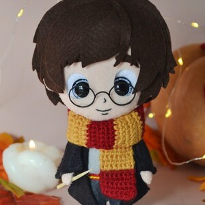 Puede incluir: Muñeca de fieltro hecha a mano de un personaje con cabello castaño, gafas redondas y una bufanda roja y dorada. La muñeca lleva una túnica negra y sostiene una varita. La muñeca es un personaje de una popular serie de fantasía.