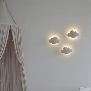 May include: Three cloud-shaped wall lights with a soft, glowing halo effect, mounted on a white wall. The lights are a light gray color and are arranged in a cluster. A canopy and crib are visible in the background.