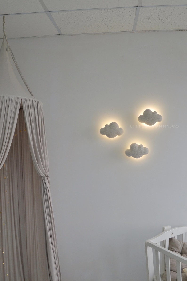 May include: Three cloud-shaped wall lights with a soft, glowing halo effect, mounted on a white wall. The lights are a light gray color and are arranged in a cluster. A canopy and crib are visible in the background.