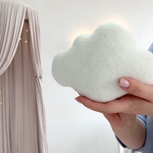 May include: A soft, white cloud-shaped pillow. The plush pillow is held in front of a white wall, with a canopy bed and string lights in the background. The pillow is a light, fluffy, and whimsical decorative item.