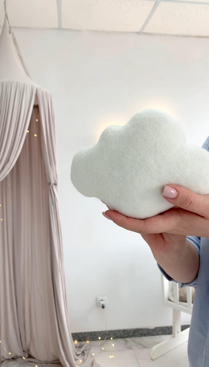 May include: A soft, white cloud-shaped pillow. The plush pillow is held in front of a white wall, with a canopy bed and string lights in the background. The pillow is a light, fluffy, and whimsical decorative item.