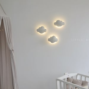 May include: Three cloud-shaped wall lights with a soft, glowing center are mounted on a white wall. The lights are part of a nursery decor, along with a white crib and a canopy. The text "LITTLE BUNNY CO" is visible on the wall.