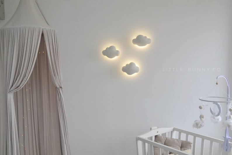 May include: Three cloud-shaped wall lights with a soft, glowing center are mounted on a white wall. The lights are part of a nursery decor, along with a white crib and a canopy. The text "LITTLE BUNNY CO" is visible on the wall.