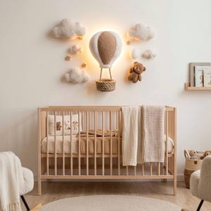 Op de afbeelding: Een kinderkamer met een houten wieg, twee crèmekleurige schommelstoelen en een aan de muur gemonteerde luchtballonlamp. De inrichting omvat wolken- en steraccenten, een teddybeer en een kleine boekenplank. Het kleurenpalet is zacht en neutraal.