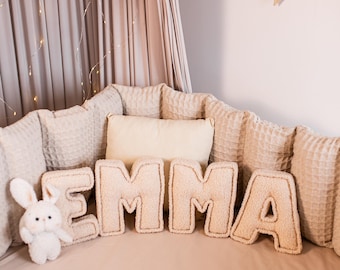 May include: A white plush bunny sits on a beige crib mattress with the name "EMMA" spelled out in large, fluffy letters. The crib is surrounded by a beige padded bumper and a canopy of sheer beige curtains.