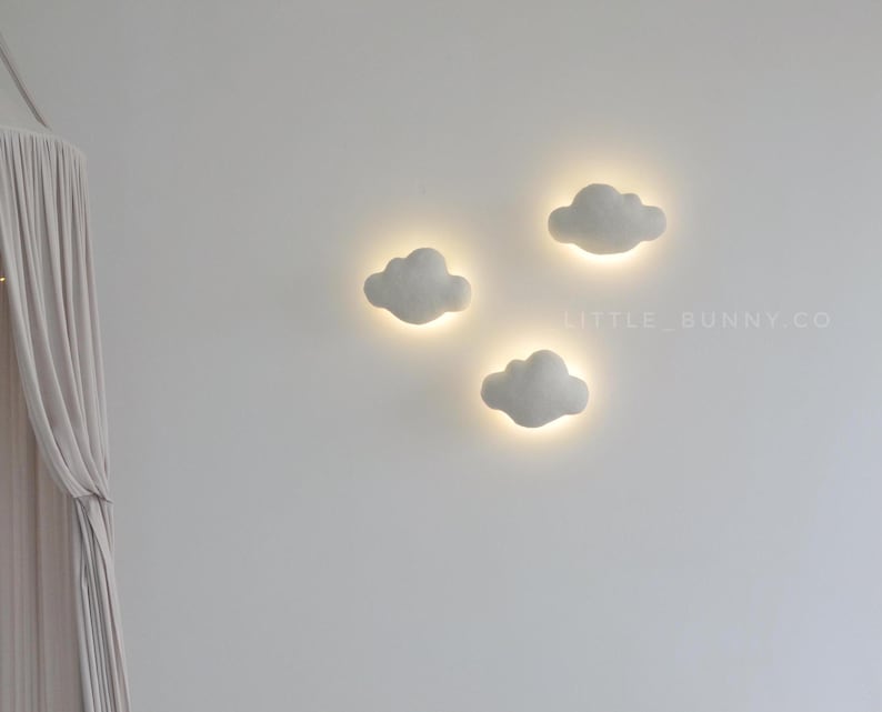 May include: Three cloud-shaped wall lights with a soft, glowing effect. The lights are a light gray color and are mounted on a white wall. The lights are arranged in a cluster, creating a whimsical and inviting atmosphere.