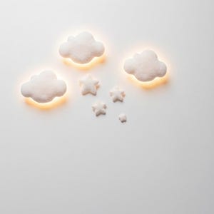 May include: White cloud and star-shaped wall lights with a warm, yellow glow. The lights are arranged in a whimsical pattern, creating a soft, ambient lighting effect. Ideal for a nursery or children's room.
