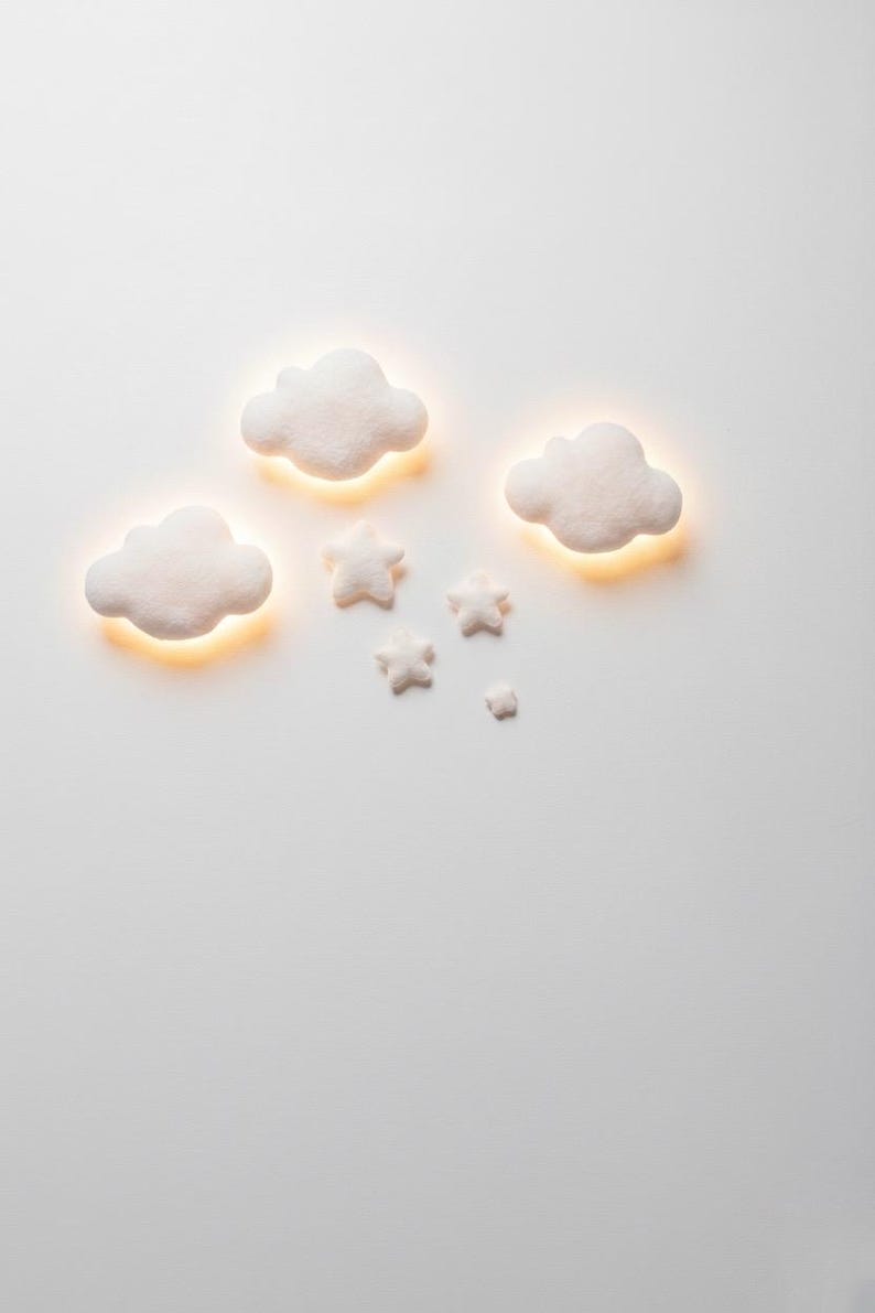 May include: White cloud and star-shaped wall decor with a warm, glowing light emanating from behind. The set includes three cloud shapes and four star shapes, creating a whimsical, dreamy aesthetic. Perfect for a nursery or children's room.