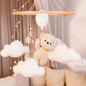 May include: A wooden baby mobile with a teddy bear, clouds, and stars hanging from it. The mobile is suspended from a wooden ring and is hanging above a white crib.