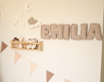May include: A nursery room decorated with a beige felt name banner that reads "EMILIA", a brown teddy bear on a cloud, a crescent moon, and stars. A wooden shelf with a small doll, a pair of baby shoes, and a wooden toy is on the wall. A pink and beige triangle banner hangs below the shelf.