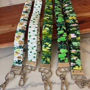Könnte beinhalten: Fünf Stoff-Lanyards mit silberfarbener Hardware. Die Lanyards weisen verschiedene grüne und weiße Muster auf, darunter Kleeblätter und florale Designs. Die Lanyards werden auf einem Holzschneidebrett präsentiert.