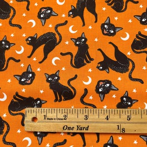 May include: Orange fabric with a repeating pattern of black cats, white stars, and white crescent moons. A ruler is placed on the fabric, showing a measurement of one yard.