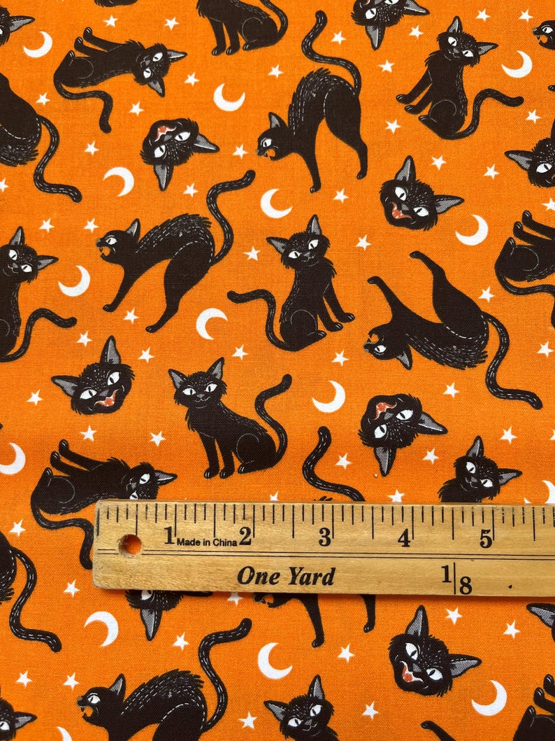 May include: Orange fabric with a repeating pattern of black cats, white stars, and white crescent moons. A ruler is placed on the fabric, showing a measurement of one yard.