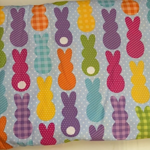 Peut inclure: Tissu bleu clair avec un motif répété de lapins de Pâques colorés. Les lapins sont de différentes couleurs, notamment rose, violet, orange, jaune, vert et bleu, avec des motifs à pois, à rayures et à carreaux.