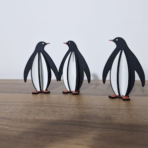 May include: Three black and white penguin figurines standing on a wooden surface. The penguins are made of wood and have a simple, modern design.