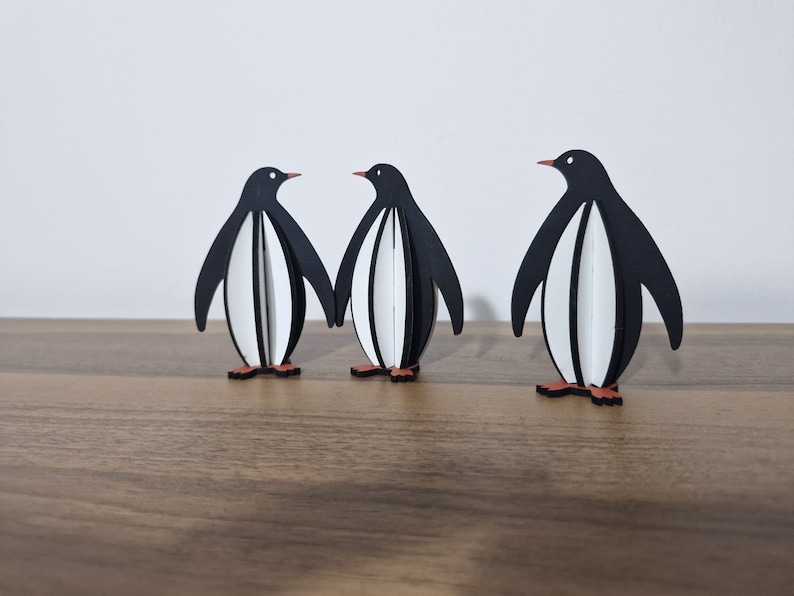 May include: Three black and white penguin figurines standing on a wooden surface. The penguins are made of wood and have a simple, modern design.