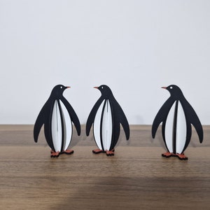 May include: Three black and white penguin figurines standing on a wooden surface. The penguins are facing forward with their wings outstretched.