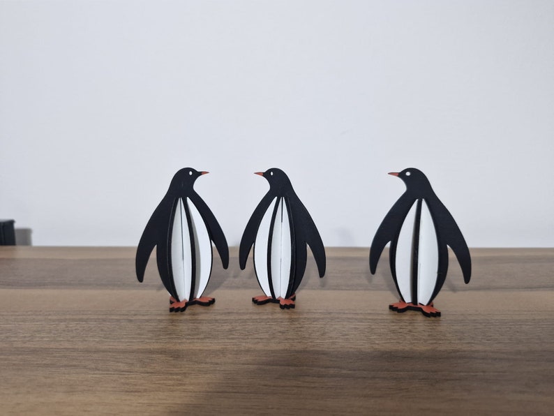 May include: Three black and white penguin figurines standing on a wooden surface. The penguins are facing forward with their wings outstretched.