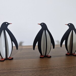 May include: Three black and white penguin figurines with orange feet. The penguins are made of wood and are standing on a wooden surface.