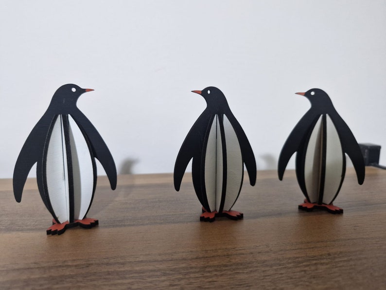 May include: Three black and white penguin figurines with orange feet. The penguins are made of wood and are standing on a wooden surface.