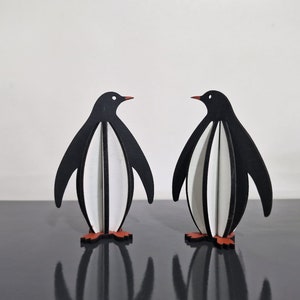 May include: Two black and white penguin figurines with red feet. The penguins are made of wood and are standing on a black surface.