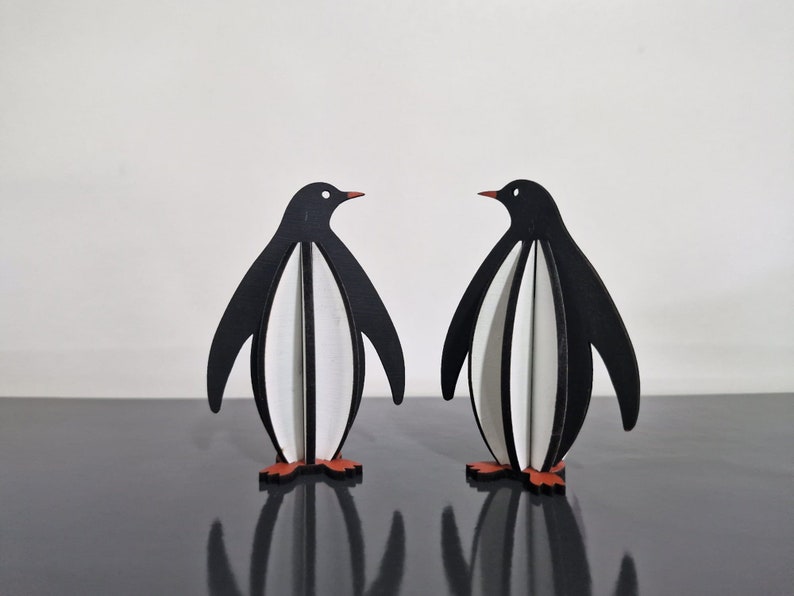 May include: Two black and white penguin figurines with red feet. The penguins are made of wood and are standing on a black surface.