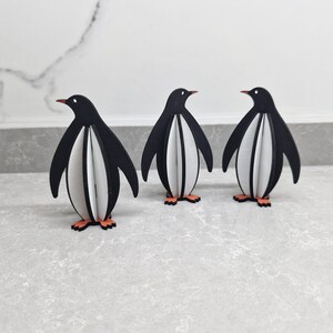 May include: Three black and white penguin figurines standing on a gray surface. The penguins are made of wood and have a 3D design.